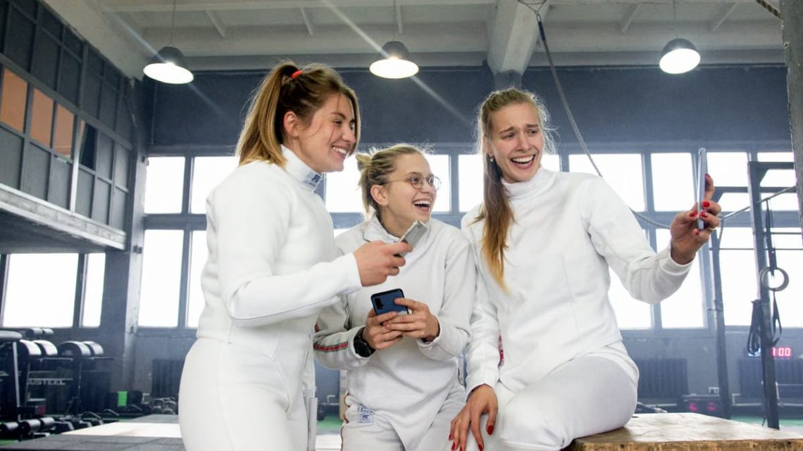 Three women fencers in white uniforms laughing and taking a selfie indoors.
