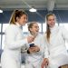 Three women fencers in white uniforms laughing and taking a selfie indoors.